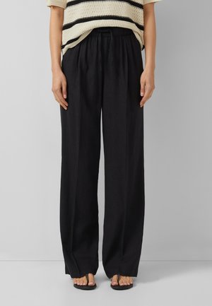 Frau trägt lockere schwarze Hosen mit weitem Bein und elastischem Bund, kombiniert mit schwarzen Sandalen und einem beige gestreiften gehäkelten Oberteil.