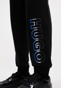 Melni sporta bikses ar lielu, atstarojošu "HUGO" logotipu metāliski sudrabā un zils gar kāju. Mīksts materiāls ar piestiprinātu piedurkni.