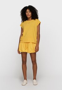 Mujer de pie, vestida con un top amarillo de manga corta y una mini falda fruncida a juego, con zapatillas blancas, sobre un fondo claro y liso.