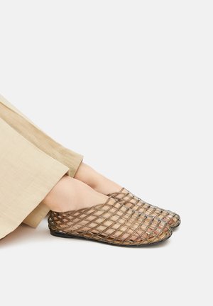 Transparent woven shoes with an open mesh design, featuring a brownish tint and a flat sole, paired with a light beige pant.