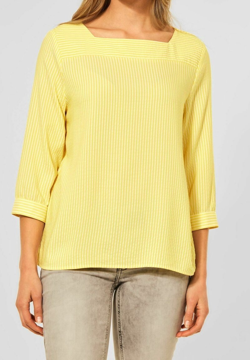 Haut jaune à manches longues avec des rayures blanches et un col carré, porté avec un jean gris clair, visible des épaules aux hanches.