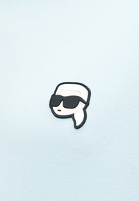 Στυλιζαρισμένο καρτούν κονκάρδα με πρόσωπο που έχει λευκά μαλλιά σε αλογοουρά και μεγάλα μαύρα γυαλιά ηλίου, σε φόντο ανοιχτό μπλε ύφασμα.