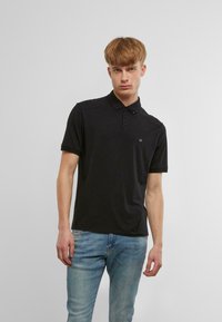 Fekete pólóing gallérral és három gombbal, a mellkason lévő kis logóval. Az anyag simának tűnik, és testhezálló fazonú.