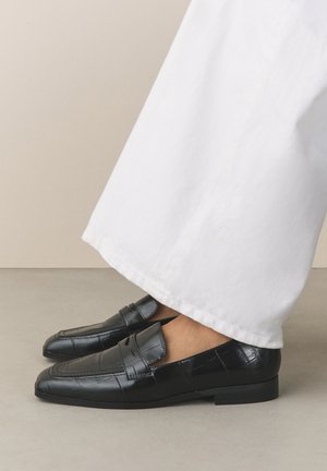 Zwarte leren loafers met een textuurpatroon gedragen met witte broek met wijde pijpen op een neutrale grijze vloer.