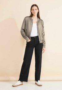Veste bomber gris clair avec une texture lisse, poches zippées, portée sur un haut blanc. Assortie avec un jean noir à taille haute et à jambes larges, ainsi que des baskets.