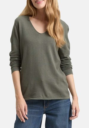 Femme portant un pull à manches longues en maille vert olive et un jean bleu, debout les bras détendus contre un fond blanc.
