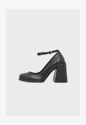 Chaussure à talons hauts en cuir noir à bout fermé avec un talon bloc épais et une bride de cheville réglable avec deux petites boucles.