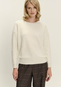 Blondynka w białym swetrze z długim rękawem i brązowych spodniach w kratę stoi na tle gładkiej ściany.