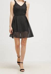 Robe noire avec un corsage ajusté et une jupe évasée, ornée de rayures horizontales et de couches transparentes. Portée avec des sandales à talons noires.