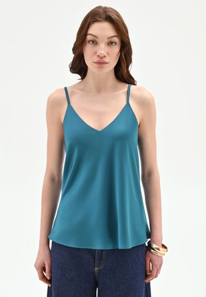 Femme portant un haut sans manches à col en V teal et un jean bleu foncé, debout avec une expression neutre et des bracelets en or à son poignet droit.