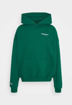 Donkergroene capuchontrui met voorzak, witte tekst "WORST BEHAVIOR" op de borst en kleine witte tekst op de linker mouwboord.