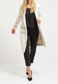 Lang, witte jas met gestreepte patronen in pastelkleuren, gedragen over een zwarte blouse en zwarte broek, met een getailleerde pasvorm en gestructureerde stof.