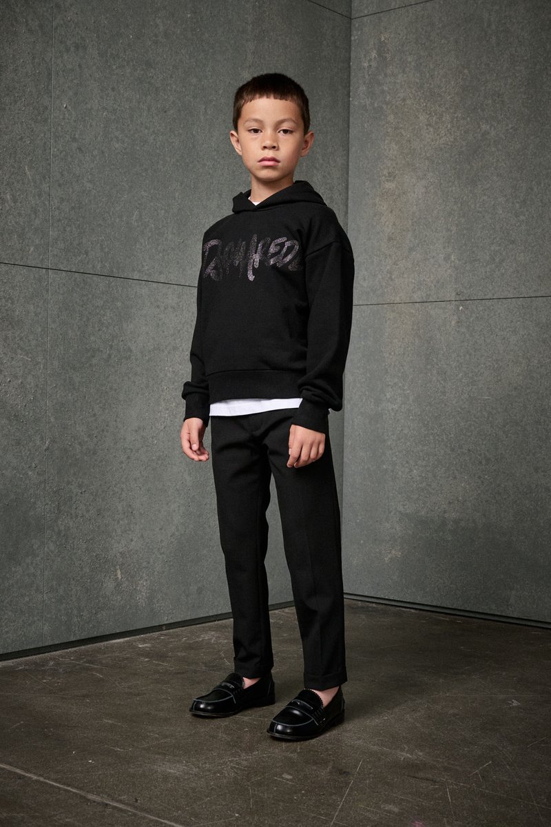 Schwarzer Hoodie mit metallischen Buchstaben, über einem weißen Shirt getragen. Schwarze maßgeschneiderte Hose und glänzende schwarze Loafers vervollständigen das Outfit.