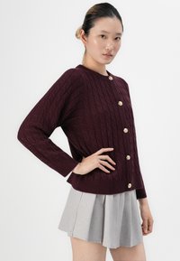 Žena v tmavě fialovém pleteném kardiganu s zlatými knoflíky a světle šedou plisovanou sukní, pózuje na bílém pozadí.