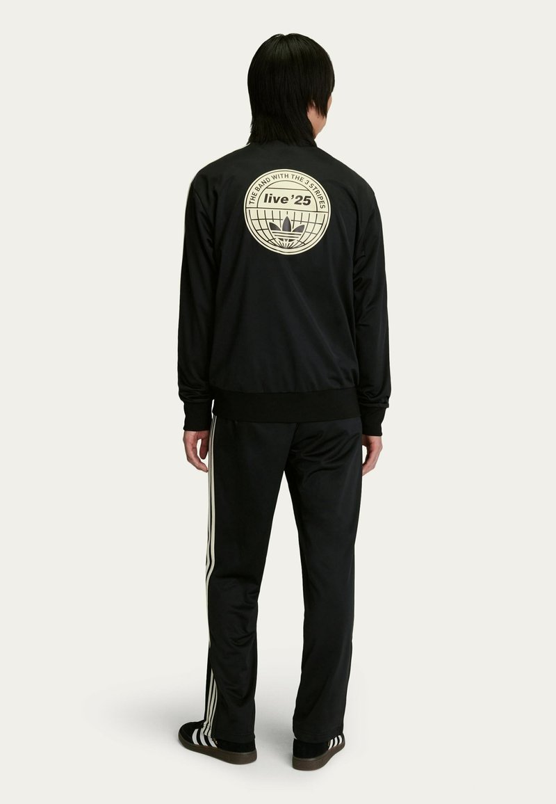 Sort trackjakke med et cremefarvet logo og grafik på bagsiden, sammen med sorte trackbukser med hvide sidelister.