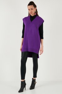 Žena u ljubičastoj tunici bez rukava s V-izrezom preko crne majice, crnim tajicama, crnim gležnjačama i velikim ružičastim naušnicama na bijeloj pozadini.