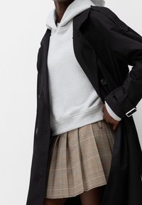 Czarny trenchcoat na szarym hoodie, w połączeniu z plisowaną spódnicą w kratkę w beżowych i brązowych odcieniach. Stylizacja prezentuje detal z guzikami na spódnicy.