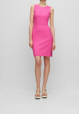 Mujer con un vestido ajustado sin mangas de color rosa brillante con una pequeña abertura lateral, combinado con sandalias de plataforma beige sobre un fondo liso.
