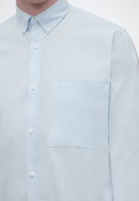Ανοιχτό μπλε πουκάμισο με κουμπιά, γιακά με κουμπιά και αριστερή τσέπη στο στήθος με διακριτικές ραφές.
