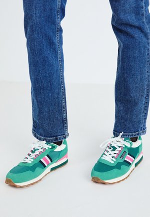 Persoon die een blauwe spijkerbroek en groene sneakers draagt met witte veters en roze, witte en zwarte strepen op een lichte achtergrond.