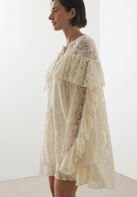 Spitzenkleid in Creme mit lockerer Passform. Verfügt über Rüschensäume, ein strukturiertes Muster und einen Bindebandverschluss am Hals.