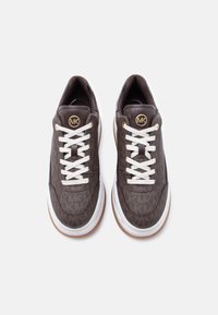 Ζεύγος καφέ αθλητικών παπουτσιών με μοτίβο, λευκά κορδόνια και χοντρές λευκές σόλες, με χρυσλό λογότυπο MK στη γλώσσα, σε θέα από ψηλά.