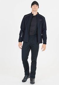 Veste de sport noire avec des accents réfléchissants sur une chemise noire, associée à un pantalon et des bottes noires, idéale pour des activités en plein air.