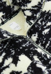 Jachetă din fleece cu fermoar, în negru și crem, cu un model abstract, având un fermoar argintiu și o căptușeală moale și texturată. Eticheta brandului este vizibilă.