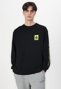 Black long-sleeve T-shirt featuring a yellow graphic on the left chest and yellow text along the right sleeve. Cotton fabric, relaxed fit.