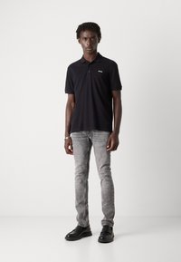 Camisa polo negra de manga corta con un pequeño logotipo blanco. Llevada con unos vaqueros gris ajustados y zapatos negros, de pie frente a un fondo liso.