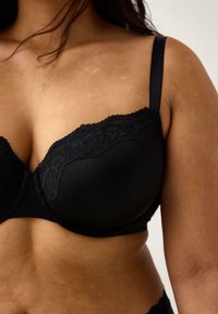 Kvindelig iført en sort bh med blonderdetaljer på skålene, der viser den øverste del af overkroppen og en del af armen mod en ensfarvet baggrund.