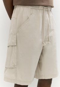 Pantaloni cargo beige con cintura elastica, chiusura con cordino, tasche laterali e tessuto texture. Presentano dettagli di cucitura visibili.