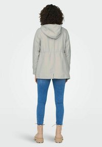 Světle šedá bunda s kapucí, elastickým pasem, zadním rozparkem a hladkou texturou, doplněná modrými úzkými džíny a béžovými kotníkovými botami.