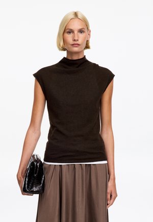 Femme blonde aux cheveux courts porte un haut noir sans manches et une jupe marron, tenant une pochette noire texturée sur fond blanc.