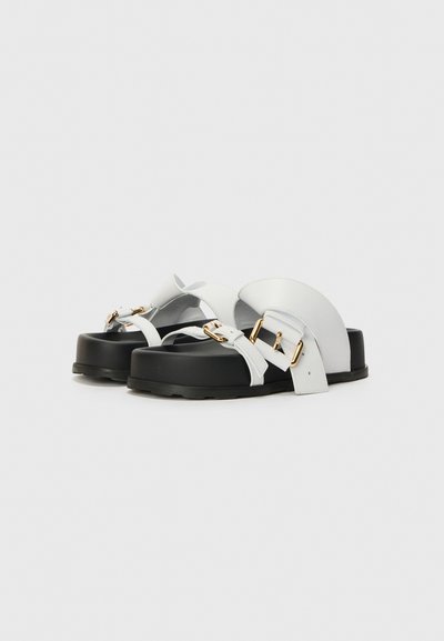 Hvide læder sandaler med sorte platform såler, med krydsede stropper og hardware i guld tone. Glat tekstur med minimale design detaljer.