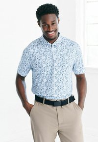 Kurzärmliges hellblaues Poloshirt mit einem sich wiederholenden Muster kleiner dunkelblauer Formen, kombiniert mit beigen Hosen und einem schwarzen Gürtel.