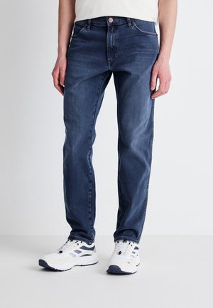 Mann in dunkelblauer Slim-Fit-Jeans und weißen Turnschuhen steht vor hellem, einfarbigem Hintergrund.