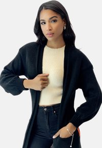 Svart stickad cardigan med lös passform, ribbade manschetter och öppen framdesign, parat med en krämfärgad ribbad topp och svarta jeans.