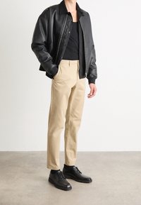 Giacca bomber in pelle nera, top nero senza maniche, pantaloni beige, scarpe stringate nere. La giacca ha una zip frontale e polsini a coste.