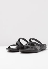 Black sandals with a soft, textured sole, featuring multiple curved straps and open sides for ventilation. Lightweight and flexible design.