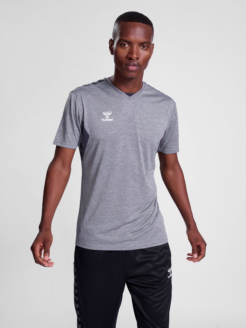Young man wearing a grey hummel sports t-shirt and black hummel sports pants, standing against a plain light background.