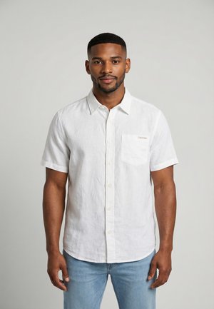 Homme aux cheveux courts portant une chemise blanche à manches courtes boutonnée et un jean bleu clair, sur un fond gris uni.