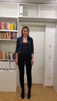 Γυναίκα με μαύρη ενδυμασία και ναυτικό σακάκι στέκεται μπροστά σε μια λευκή πόρτα και βιβλιοθήκη με βιβλία και κουτιά φακέλων σε ένα δωμάτιο με ξύλινο πάτωμα.