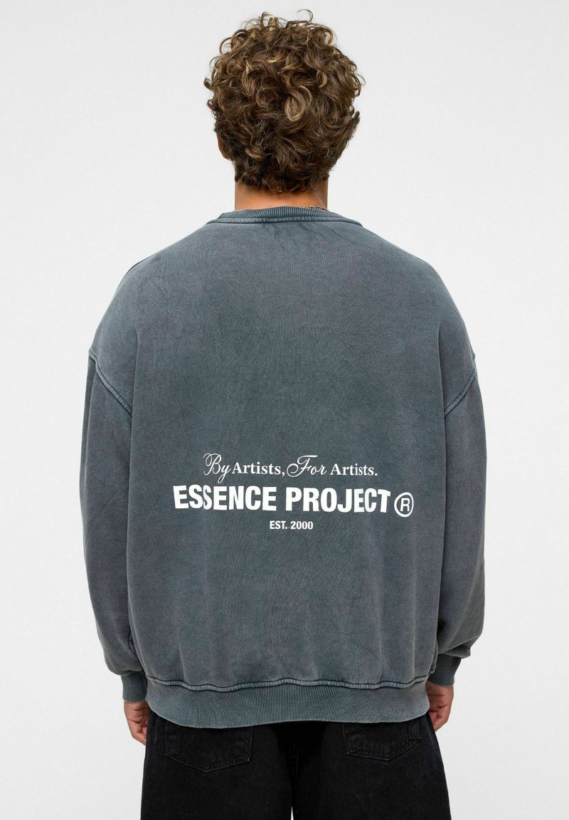 Grijze oversized sweatshirt met een ronde hals, met op de achterkant witte tekst: "Door kunstenaars, Voor kunstenaars. ESSENCE PROJECT ® OPGERICHT. 2000."