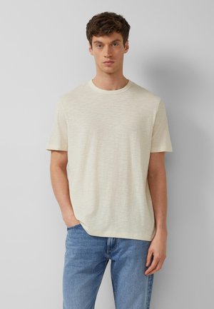 Młody mężczyzna z krótkimi brązowymi włosami, ubrany w gładką jasnobeżową koszulkę z krótkim rękawem i niebieskie dżinsy, stojący na szarym tle.