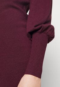 Maglione borgogna lavorato a maglia con texture a coste, caratterizzato da maniche a sbuffo e polsini aderenti, che presenta una finitura liscia e un pattern di punto uniforme.