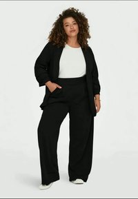 Musta ylisuuria bleisereitä valkoisen paidan päällä, yhdistetty leveälahkeisiin mustiin housuihin ja valkoisiin sneakerseihin; sileä kangas rentoon istuvuuteen.