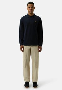 Marineblaues Langarm-Polohemd mit Kragen und einem Logo auf der Brust, kombiniert mit beigen Cargo-Hosen mit Seitentaschen und schwarzen Sneakern.