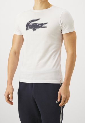 Mann trägt ein weißes T-Shirt mit großem dunklem Krokodil-Logo auf der Brust und schwarze Hose mit Logostreifen an der Seite.