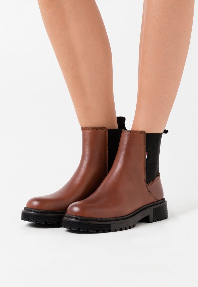 Bottes en cuir marron montant à la cheville avec des semelles noires épaisses portées sur des jambes nues sur un fond blanc.